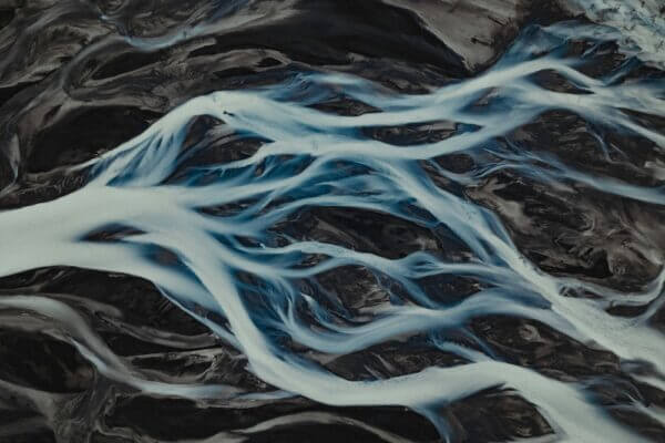 Is God Everywhere? The Opposite is Too. ∴ WholeSpirit Is God Everywhere? The opposite is too. Aerial view of dark volcanic terrain braided by bright blue glacial rivers. ∴ WholeSpirit